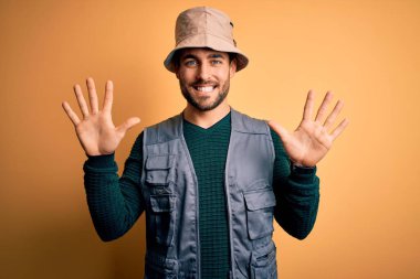 Tatilde sakallı yakışıklı turist sarı arka planda kaşif şapkası takıyor ve parmaklarını on numarayı gösterirken kendinden emin ve mutlu bir şekilde gülümsüyor..