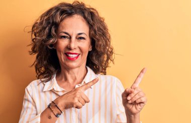 Orta yaşlı, güzel, esmer, çizgili gömlek giyen, sarı arka planda duran, gülümseyen ve kameraya bakan, iki eliyle ve parmaklarıyla yüzünü gösteren bir kadın..