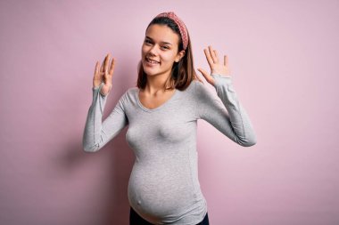 Genç, güzel bir kız hamile, pembe arka planda bebek bekliyor. Dokuz numaralı parmaklarıyla işaret ediyor. Kendinden emin ve mutlu bir şekilde gülümsüyor..