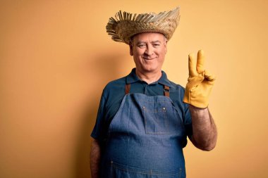 İzole edilmiş sarı arka planda önlük ve şapka takan orta yaşlı çiftçi kameraya gülümseyip zafer işareti yapıyor. İki numara..