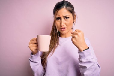 Genç, güzel, esmer bir kadın izole edilmiş pembe arka planda bir fincan kahve içiyor. Öfkeyle bağırıyor, çıldırıyor ve öfkeyle bağırıyor.