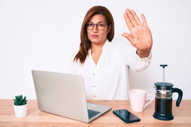 Orta yaşlı esmer, İspanyol iş kadını ofiste çalışıyor. Eli açık, dur işareti yapıyor. Ciddi ve kendinden emin bir ifadeyle. 