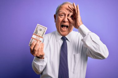 Mor arka planda elli dolarlık banknotlar tutan yaşlı gri saçlı adam gülümseyen yüzüyle tamam imzasını atarken parmaklarının arasından bakıyor.