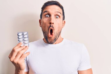 Elinde ilaç hapları olan yakışıklı bir genç beyaz arka planda korkmuş ve ağzı açık bir şekilde şaşkınlık içinde bekliyor.