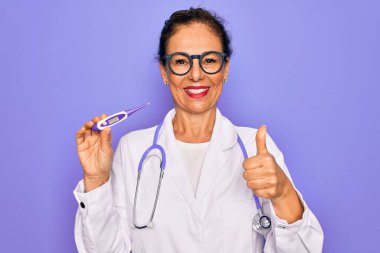 Orta yaşlı profesyonel doktor kadın elinde medikal termometre tutuyor. Gülümsüyor. Tamam işareti yapıyor.