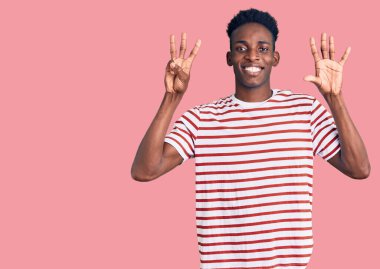 Genç, Afro-Amerikalı, günlük giysiler giymiş, 8 numaralı parmaklarıyla işaret ederken, kendinden emin ve mutlu bir şekilde gülümsüyor.. 