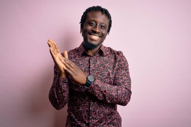Genç, yakışıklı, Afro-Amerikan bir adam, günlük gömleğini giymiş pembe arka planda duruyor, alkışlıyor ve mutlu, neşeli, gururlu elleri alkışlıyor.