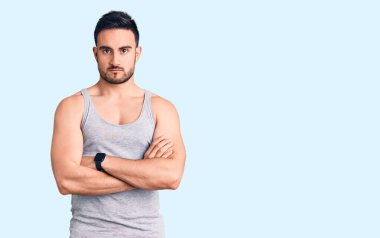 Mayo ve kolsuz tişört giyen genç yakışıklı adam şüpheci ve gergin, yüzünde çapraz kollu tasvip etmeyen bir ifade var. negatif kişi. 