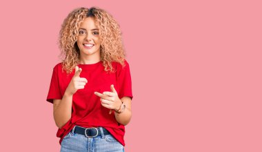 Kıvırcık saçlı, sıradan kırmızı tişörtlü, mutlu ve komik yüzlü genç sarışın kadın kamerayı işaret ediyor. iyi enerji ve titreşimler. 