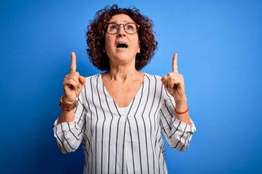 Orta yaşlı, kıvırcık saçlı, sıradan çizgili gömlek giyen güzel bir kadın izole arka planda şaşkın şaşkın bakıyor, parmaklarıyla işaret ediyor ve kollarını kaldırıyordu..