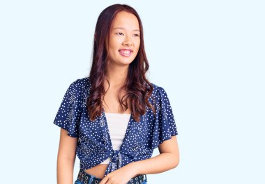 Genç, güzel Çinli bir kız, günlük kıyafetler giyip, yüzünde gülümsemeyle yan yana bakıyor, doğal bir ifade. Kendinden emin gülen. 