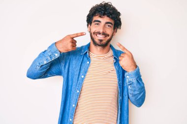 Kıvırcık saçlı, sıradan kot ceket giyen, neşeli gülümseyen ve parmaklarını, dişlerini ve ağzını gösteren yakışıklı bir genç. diş sağlığı kavramı. 