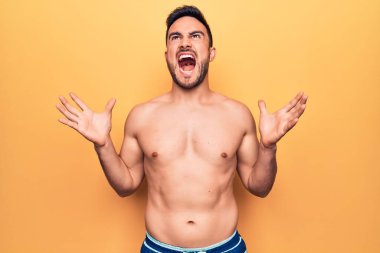 Sakallı, kolsuz tişörtlü genç bir adam sarı arka planda dikiliyor deli gibi bağırıyor ve agresif bir ifadeyle bağırıyor ve kollarını kaldırıyor. Hayal kırıklığı konsepti.