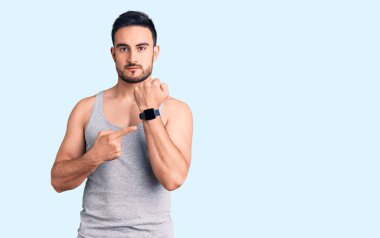 Mayolu ve kolsuz tişörtlü genç bir adam saati, sabırsızlığı ve rahat bir ifadeyle kameraya bakmayı işaret ediyor. 