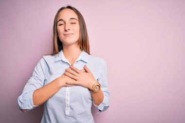 Zarif bir gömlek giyen genç ve güzel bir iş kadını soyutlanmış pembe arka planda duruyor. Elleri göğsünde, gözleri kapalı ve yüzünde minnet dolu bir jest ile gülümsüyor. Sağlık kavramı.