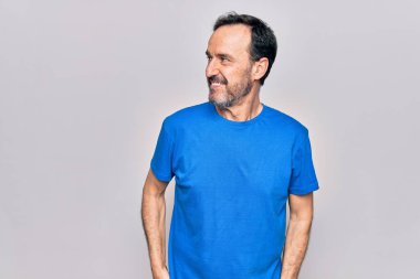 Orta yaşlı yakışıklı bir adam günlük tişört giyip izole edilmiş beyaz arka planda duruyor. Yan tarafa bakıyor, rahat bir görünüm takınıyor ve kendine güvenen bir gülümseme sergiliyor..