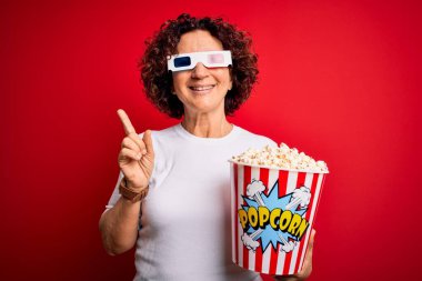 Orta yaşlı kıvırcık saçlı kadın film izlerken 3D gözlük takıyor patlamış mısır yiyor.
