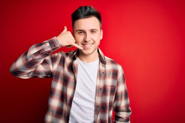 Kırmızı izole edilmiş arka plan üzerine gündelik modern tişört giyen genç beyaz adam telefonla el ve parmaklarıyla telefonla konuşuyor gibi gülümsüyor. İletişim kavramları.