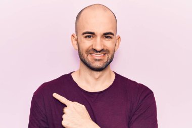 Genç, yakışıklı, kel bir adam, günlük tişört giyiyor. Gülümsüyor, eli ve parmağı yana doğru uzanıyor. 