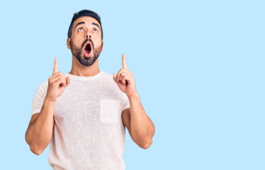 Gündelik giysiler giyen genç İspanyol adam şaşırmış ve şaşırmış bir şekilde yukarı bakıyor ve parmaklarıyla işaret ediyor ve kollarını kaldırıyor.. 