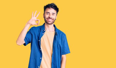 Gündelik kıyafetler giyen genç İspanyol adam gülümsüyor. El ve parmaklarıyla 