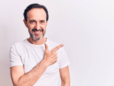 Orta yaşlı yakışıklı bir adam, günlük tişört giyiyor, izole edilmiş beyaz arka planda duruyor, gülümsüyor, eli ve parmağı yana doğru uzanıyor.