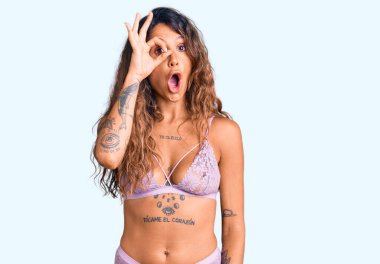 Dövmeli, iç çamaşırlı genç İspanyol kadın şaşırmış yüz ifadesiyle şaşırmış, parmaklarının arasından bakan bir jest yapıyor. İnançsız ifade. 