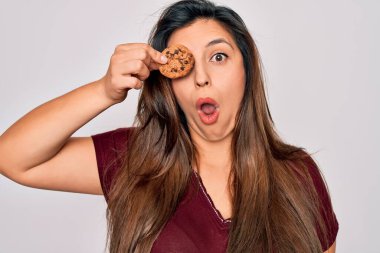 Young hispanic woman eating sweet chocolated chips cookie over isolated background scared in shock with a surprise face, afraid and excited with fear expression clipart