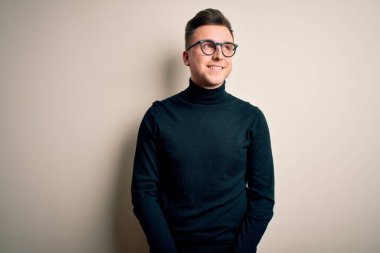 Gözlük takan yakışıklı beyaz bir adam izole edilmiş arka planda rahat bir süveter giyiyor. Yüzünde gülümseme, doğal bir ifade ile yan yana bakıyor. Kendinden emin gülen.