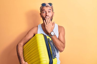 Güneş gözlüğü takan genç, sarışın, tatilde olan yakışıklı bir turist. Elinde uçak kabini çantası var. Ağzını eliyle kapatıyor. Şaşırtıcı ifade.