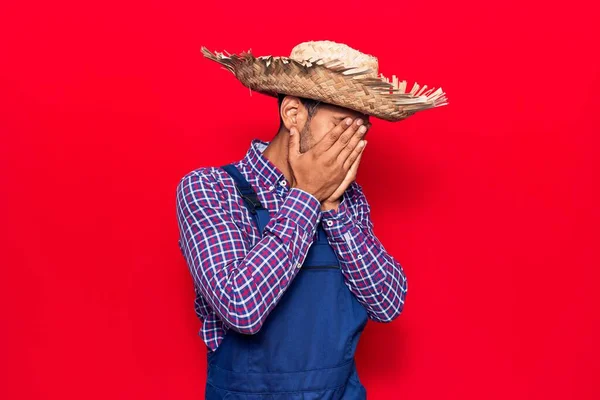 Young latin man wearing farmer hat and apron with sad expression covering face with hands while crying. depression concept.
