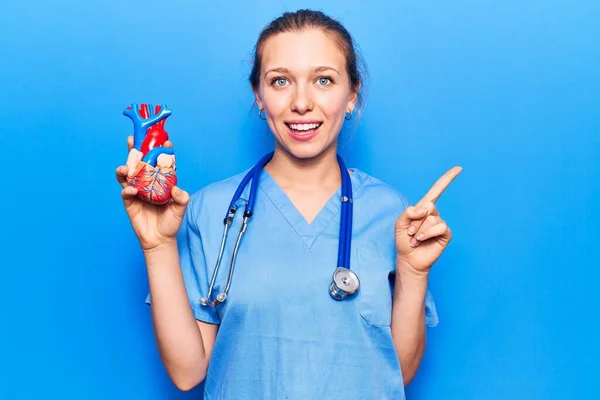 Genç Sarışın Kadın Doktor Üniforması Giyiyor Kalbi Tutuyor Gülümsüyor — Stok fotoğraf