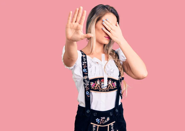 Young beautiful blonde woman wearing oktoberfest dress covering eyes with hands and doing stop gesture with sad and fear expression. embarrassed and negative concept.