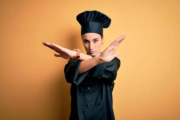 Young beautiful chef woman wearing cooker uniform and hat standing over yellow background Rejection expression crossing arms and palms doing negative sign, angry face