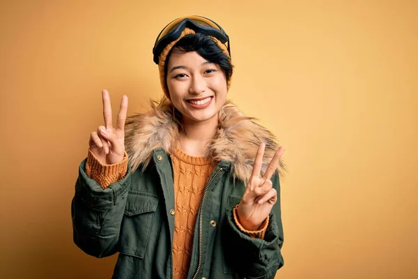 黄色の背景にスキーゴーグルを使用して雪のスポーツウェアを身に着けている美しいアジアのスキー少女は 勝利のサインを行う指を示すカメラを見て微笑んだ 第二番 — ストック写真