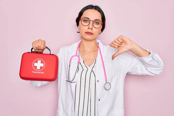 Young beautiful doctor woman with blue eyes wearing stethoscope holding first aid kit box with angry face, negative sign showing dislike with thumbs down, rejection concept