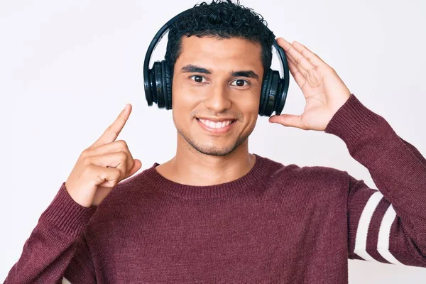 若いですハンサムなヒスパニック系の男は 側に手と指で幸せなポインティングを笑顔ヘッドフォンを使用して音楽を聴く — ストック写真