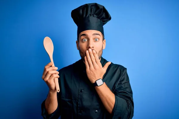年轻的厨师 留着胡子 身穿制服 用木勺捂住蓝色背景的嘴 手被错误 恐惧的表情 沉默中的恐惧 神秘的概念所震惊 — 图库照片