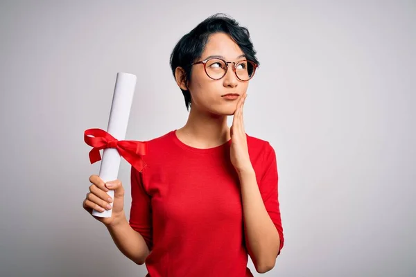 Beautiful chinese student woman wearing glasses holding university graduated diploma degree serious face thinking about question, very confused idea