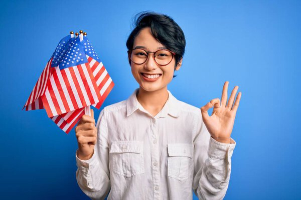 Young beautiful asian patriotic girl holding united states flags celebrating independence day doing ok sign with fingers, excellent symbol