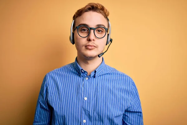 顔に真剣な表情でヘッドセットを使用して作業眼鏡をかけて働く若いハンサムな赤毛のコールセンターエージェントの男 シンプルで自然なカメラを見て — ストック写真