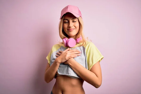 Young beautiful blonde sportswoman doing sport using headphones over pink background smiling with hands on chest with closed eyes and grateful gesture on face. Health concept.