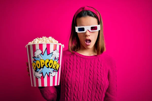 Young beautiful girl watching movie using 3d glasses eating box with popcorns In shock face, looking skeptical and sarcastic, surprised with open mouth