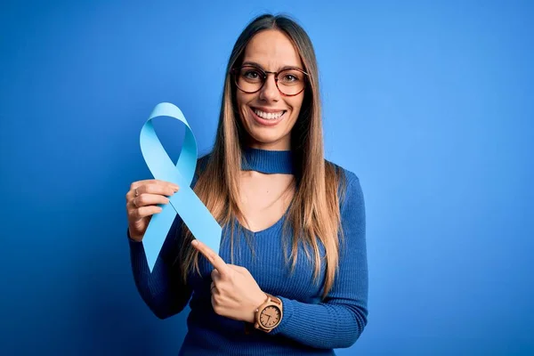 Young blonde woman with blue eyes holding colon cancer awareness blue ribbon very happy pointing with hand and finger