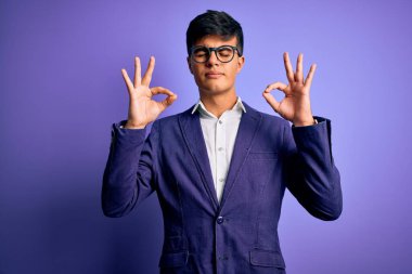 Ceket ve gözlük takan genç yakışıklı iş adamı, izole edilmiş mor arka planda rahatlamış ve parmaklarıyla meditasyon hareketi yaparken gözleri kapalı gülümsüyor. Yoga konsepti.