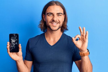 Kırık akıllı telefon tutan genç yakışıklı adam kırık ekranı gösteriyor tamam işareti yapıyor parmaklarıyla, gülümseyerek mükemmel bir sembol oluşturuyor. 