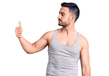 Mayo ve kolsuz tişört giyen genç yakışıklı adam gururlu görünüyor, gülümsüyor, baş parmak işareti yapıyor. 