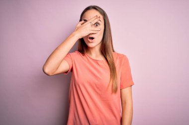 Gündelik tişört giyen genç ve güzel bir kadın izole edilmiş pembe arka planda dikiliyor. Şoka giriyor, yüzünü ve gözlerini eliyle kapatıyor ve utanmış bir ifadeyle parmaklarının arasından bakıyor..