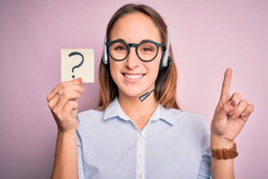Güzel çağrı merkezi ajanı kadın elinde kulaklıkla çalışıyor. Soru işareti var. Bir fikir ya da soru işareti, mutlu yüzle işaret ediyor.