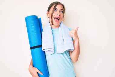 Güzel, beyaz bir kadın yoga minderini tutarken baş parmağını yukarı kaldırıyor ve ağzı açık gülümsüyor. 